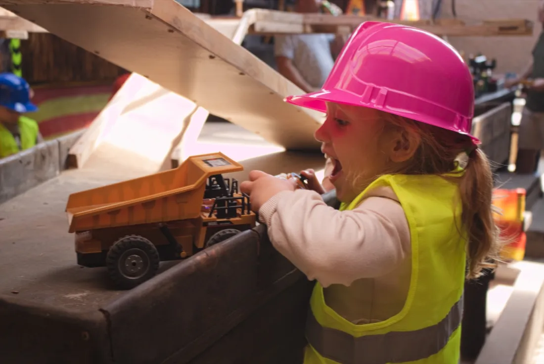 A delighted child in a pink hard hat driving a remote-control dump truck at Backyard Diggers
